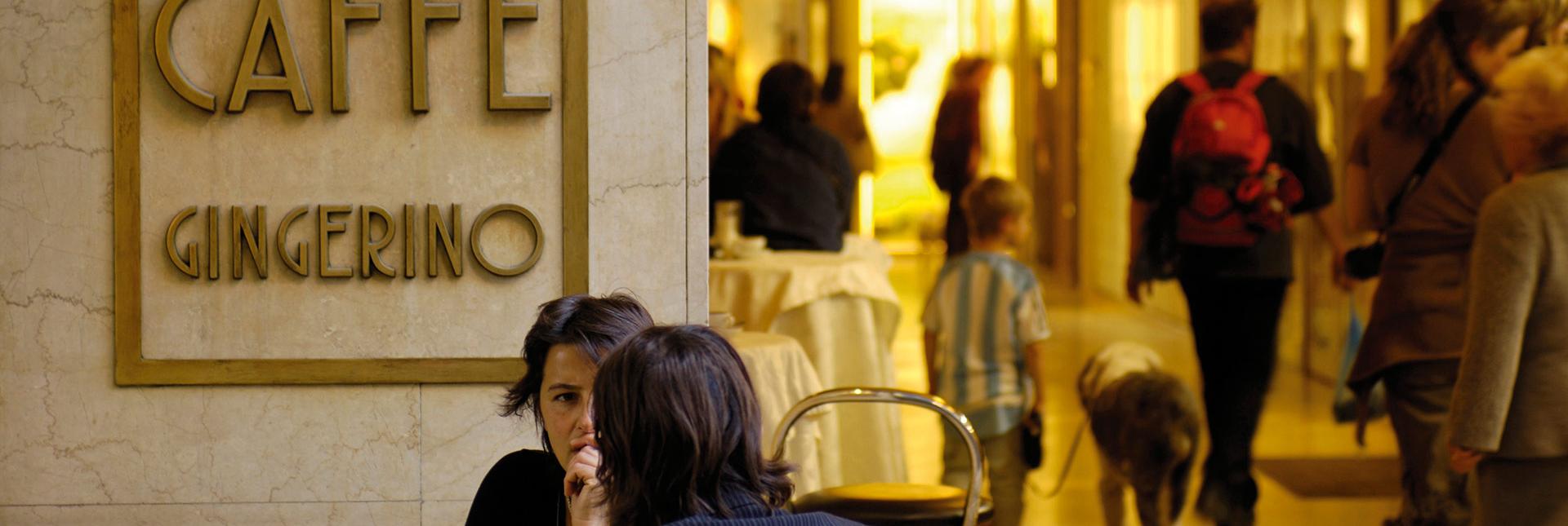 Persone parlano davanti al Caffè Gingerino a Bolzano, sullo sfondo passanti sotto i portici.